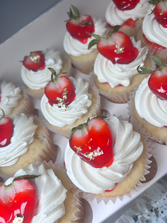 A tray of gourmet strawberry cheesecake cupcakes with white frosting and a strawberry on top, with different flavors available.